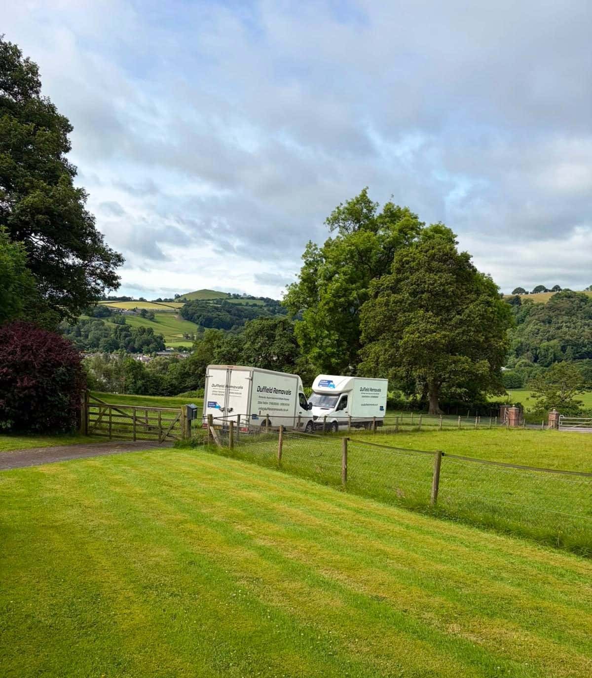 Two Duffield Removals vans by a five-bar gate in rolling green hills
