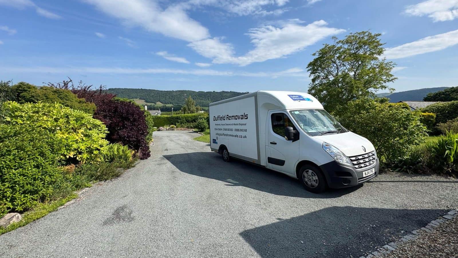 Duffield Removals Luton van on a country drive between gardens with wooded hills behind